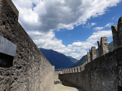 A stone walkway within a medieval fortress featuring high walls, crenellated battlements on the right, and an informational sign on the left wall, with mountains visible in the background.