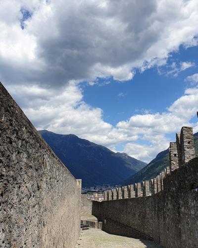 A stone walkway within a medieval fortress featuring high walls, crenellated battlements on the right, and an informational sign on the left wall, with mountains visible in the background.