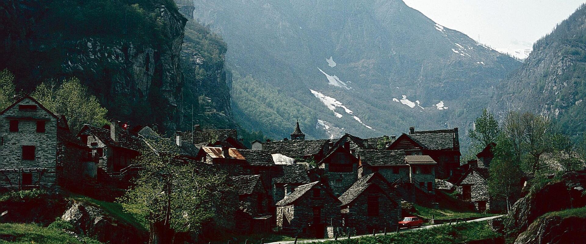 A cluster of traditional stone houses with slate roofs situated in a green valley floor, backed by steep, rugged mountains with patches of snow, and a road winding through the foreground.