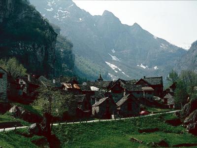 A cluster of traditional stone houses with slate roofs situated in a green valley floor, backed by steep, rugged mountains with patches of snow, and a road winding through the foreground.