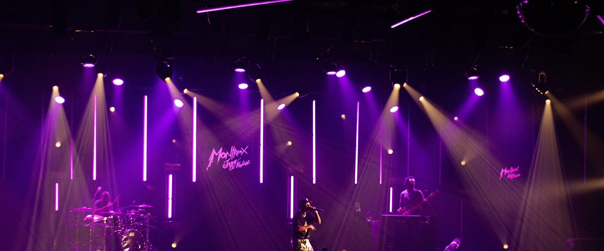 A live music performance on an indoor stage with purple lighting, featuring a band and a large neon sign in the background reading "Montreux Jazz Festival".