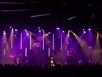 A live music performance on an indoor stage with purple lighting, featuring a band and a large neon sign in the background reading "Montreux Jazz Festival".