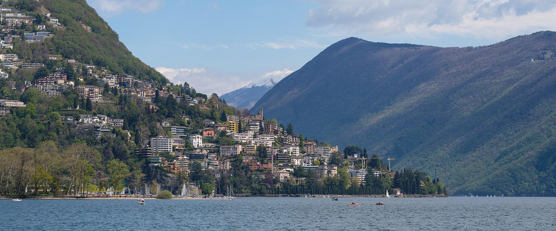 A wide landscape view of a lakeside town built on a steep hillside, with a large mountain rising behind it and snow-capped peaks visible in the distance.