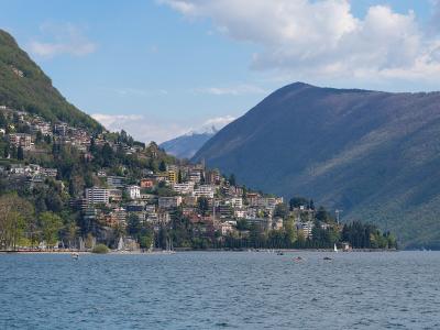 A wide landscape view of a lakeside town built on a steep hillside, with a large mountain rising behind it and snow-capped peaks visible in the distance.