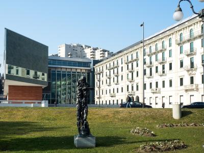 A modern building with a large angled grey facade and glass front stands next to a classic white multi-story building, with a dark abstract sculpture on a grassy lawn in the foreground.