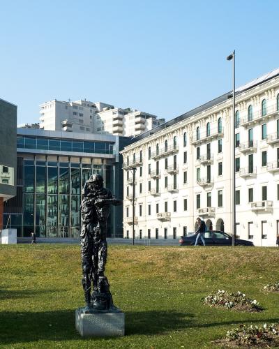 A modern building with a large angled grey facade and glass front stands next to a classic white multi-story building, with a dark abstract sculpture on a grassy lawn in the foreground.