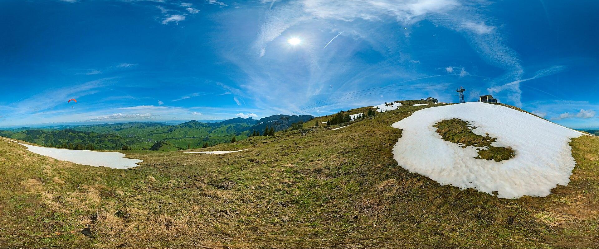 A 360-degree panoramic photograph of a grassy alpine ridge with lingering snow patches, a paraglider in the distance, and a small structure or tower on the summit against a blue sky.