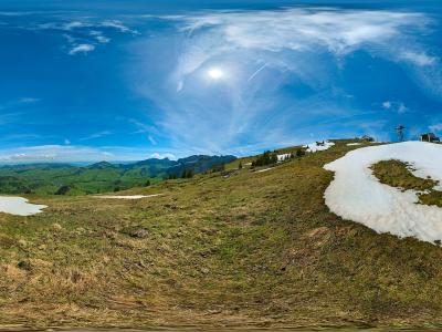 A 360-degree panoramic photograph of a grassy alpine ridge with lingering snow patches, a paraglider in the distance, and a small structure or tower on the summit against a blue sky.