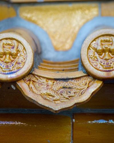 A close-up of ornate, glazed ceramic roof tiles featuring traditional Chinese dragon motifs and circular end-caps with stylized animal faces, set against a yellow and blue tiled background.