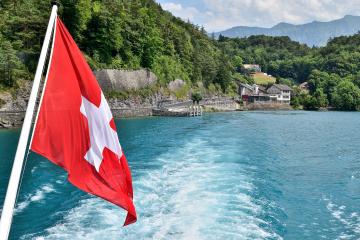 A view from the stern of a moving boat on a lake, featuring a large Swiss flag in the foreground and a lakeside building with a dock on the shore.