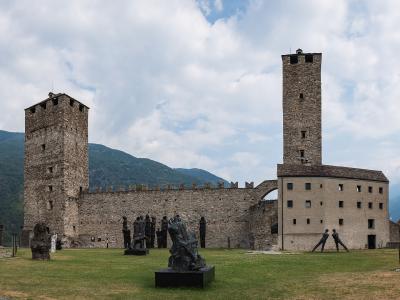 A wide shot of a medieval stone fortress complex featuring two prominent square towers and a connecting curtain wall, set on a grassy courtyard with several modern bronze sculptures in the foreground and mountains in the background.