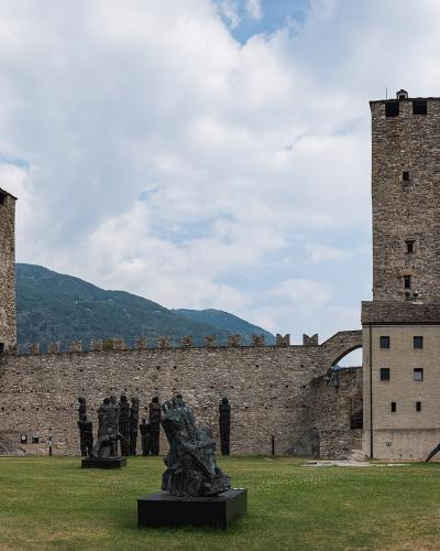 A wide shot of a medieval stone fortress complex featuring two prominent square towers and a connecting curtain wall, set on a grassy courtyard with several modern bronze sculptures in the foreground and mountains in the background.