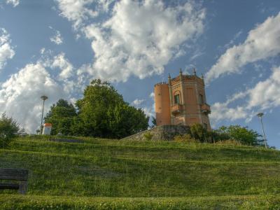 A low-angle shot of a grassy hillside leading up to a distinctive pinkish-orange, castle-like tower with crenellations and turrets, set against a blue sky with white clouds.