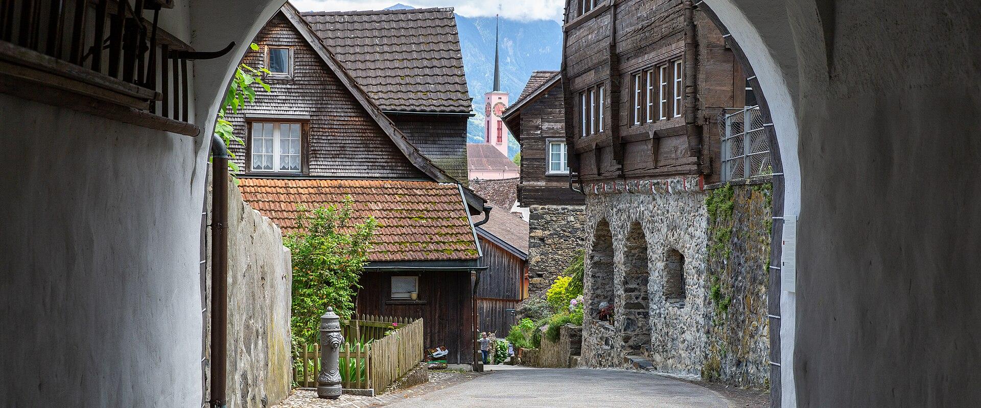 A view through a white stone archway looking onto a paved street in a historic village, featuring traditional wooden houses, a church spire in the distance, and mountains in the background.