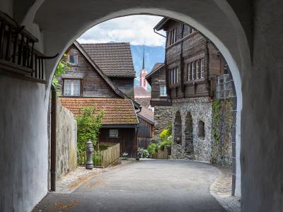 A view through a white stone archway looking onto a paved street in a historic village, featuring traditional wooden houses, a church spire in the distance, and mountains in the background.