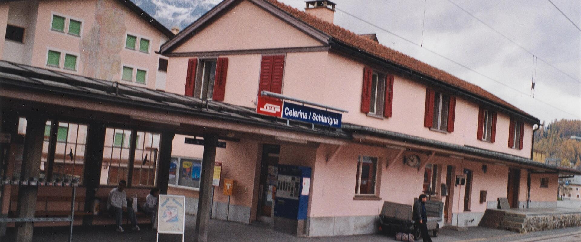 A pink station building with red shutters and a platform sign reading "Celerina / Schlariogna" under an overcast sky with mountains in the background.