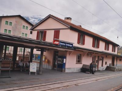 A pink station building with red shutters and a platform sign reading "Celerina / Schlariogna" under an overcast sky with mountains in the background.