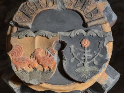 A close-up photograph of a circular, terracotta-colored architectural keystone featuring a split shield with a rooster on the left and a flower on the right, topped with the Latin inscription "DESIDERIUS MURER".