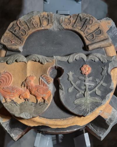 A close-up photograph of a circular, terracotta-colored architectural keystone featuring a split shield with a rooster on the left and a flower on the right, topped with the Latin inscription "DESIDERIUS MURER".