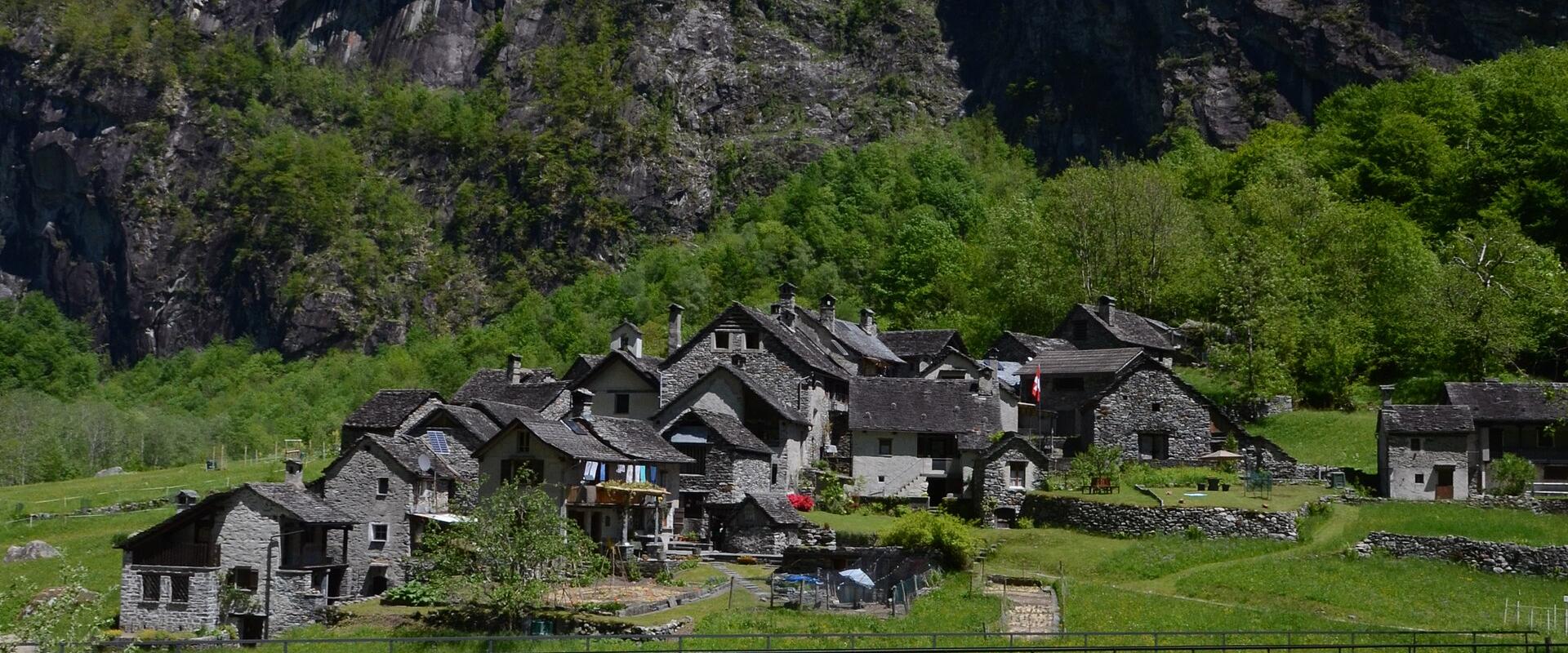 A cluster of traditional stone houses in a mountain valley with a river and bridge in the foreground, set against steep rocky cliffs.