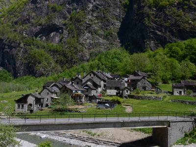 A cluster of traditional stone houses in a mountain valley with a river and bridge in the foreground, set against steep rocky cliffs.