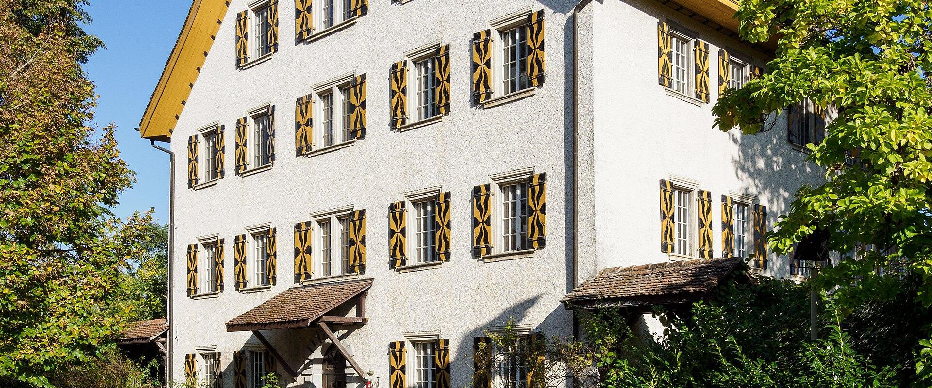 A white, multi-story building with a steep gabled roof and distinctive yellow-and-black patterned shutters, set against a blue sky with surrounding trees and a gravel path.