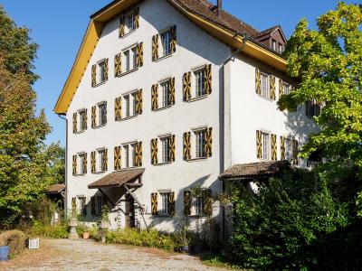 A white, multi-story building with a steep gabled roof and distinctive yellow-and-black patterned shutters, set against a blue sky with surrounding trees and a gravel path.