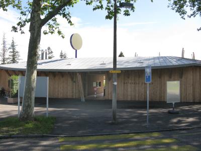 A low, single-story building with a curved wooden facade and a metal roof, featuring a circular sign on a pole and a blue pedestrian crossing sign in the foreground.