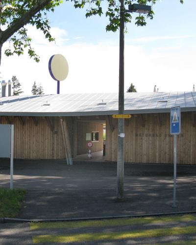 A low, single-story building with a curved wooden facade and a metal roof, featuring a circular sign on a pole and a blue pedestrian crossing sign in the foreground.