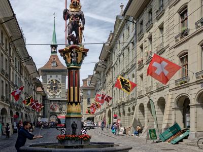 A stone fountain with a statue of a knight holding a bear and a flag, situated in a cobblestone street with Swiss flags and a clock tower in the background.