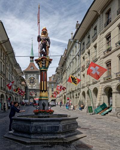 A stone fountain with a statue of a knight holding a bear and a flag, situated in a cobblestone street with Swiss flags and a clock tower in the background.