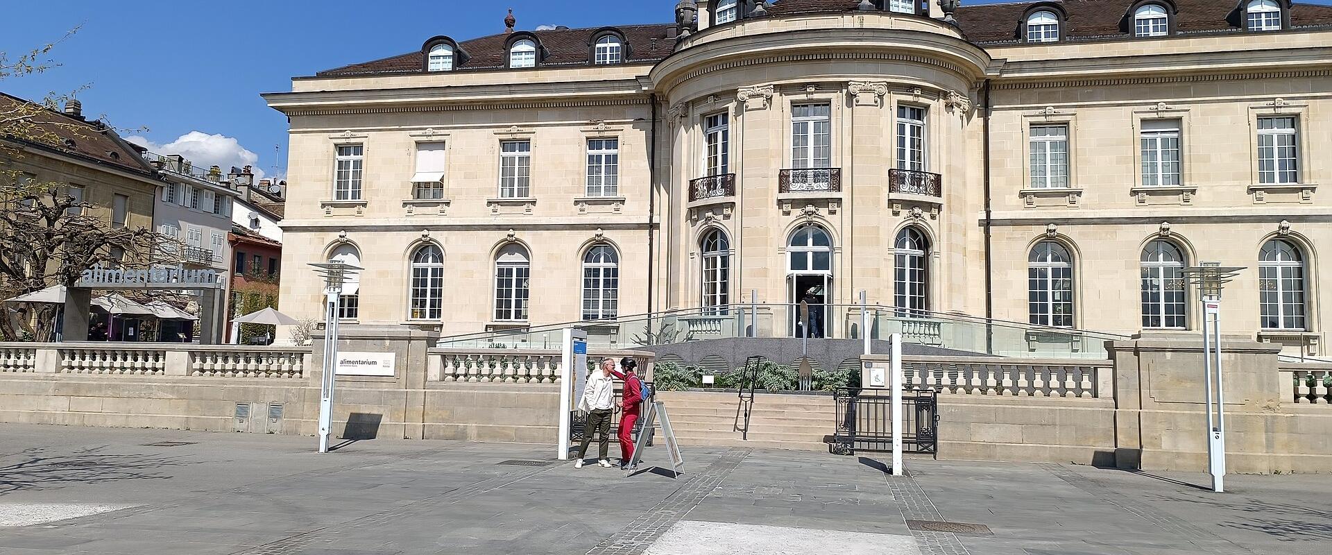A large, light-colored stone building with classical architecture featuring a prominent curved central section and mansard roof, with a sign reading "alimentarium" visible on a low wall in the foreground.
