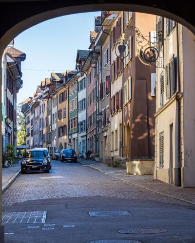 A view looking out from under a dark stone archway onto a narrow, cobblestone street lined with colorful, multi-story historic buildings and parked cars.