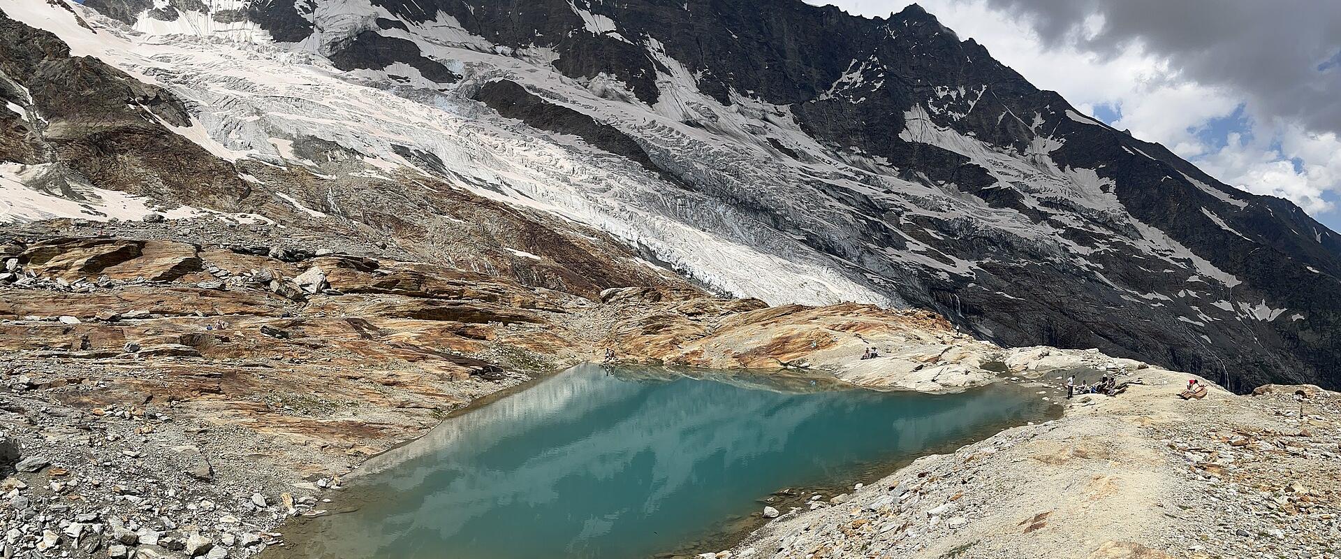 A high-altitude alpine landscape featuring a turquoise glacial lake in the foreground and a large, snow-covered glacier descending a rugged mountain face in the background.