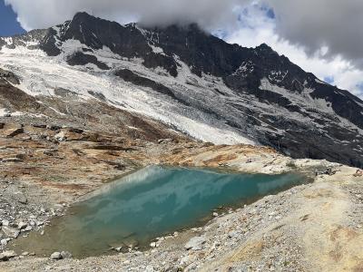 A high-altitude alpine landscape featuring a turquoise glacial lake in the foreground and a large, snow-covered glacier descending a rugged mountain face in the background.