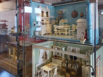 A glass display case containing two levels of antique dollhouse furniture and miniature room settings, including a kitchen and dining area with white wooden furniture and blue walls.