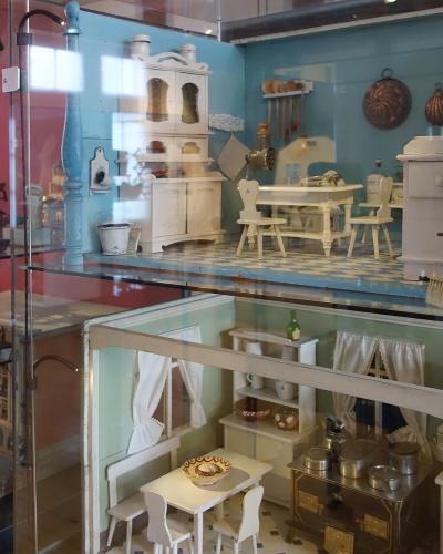 A glass display case containing two levels of antique dollhouse furniture and miniature room settings, including a kitchen and dining area with white wooden furniture and blue walls.