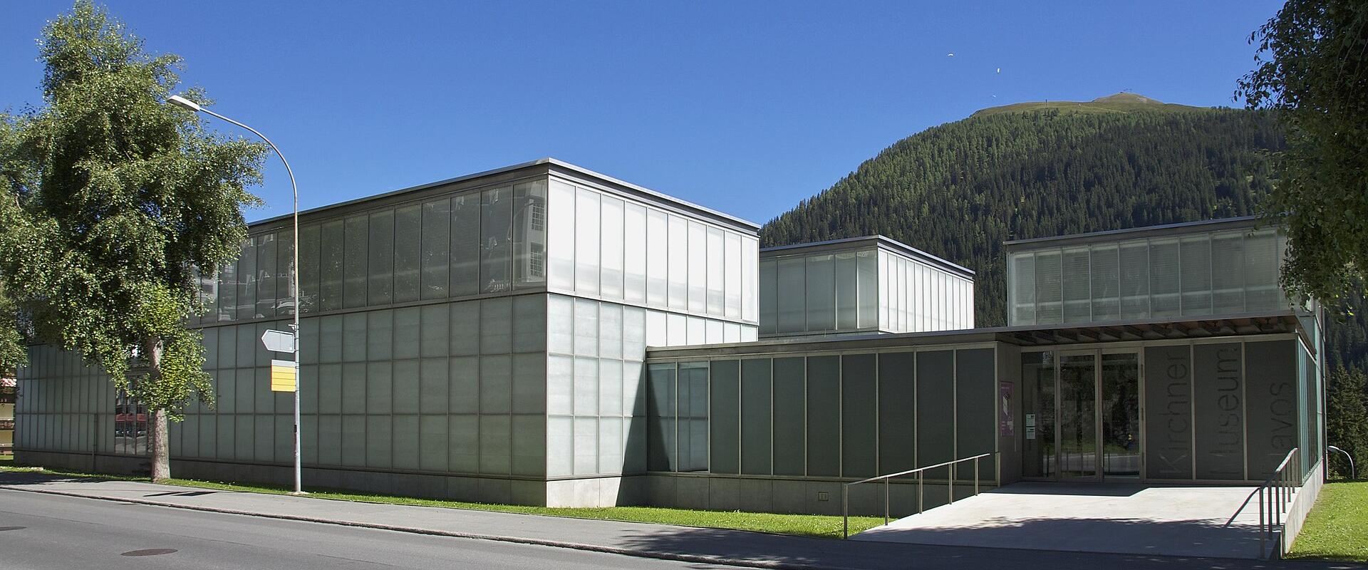 A modern, low-rise building with a glass and metal facade, situated next to a road with a forested mountain in the background.