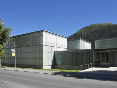 A modern, low-rise building with a glass and metal facade, situated next to a road with a forested mountain in the background.