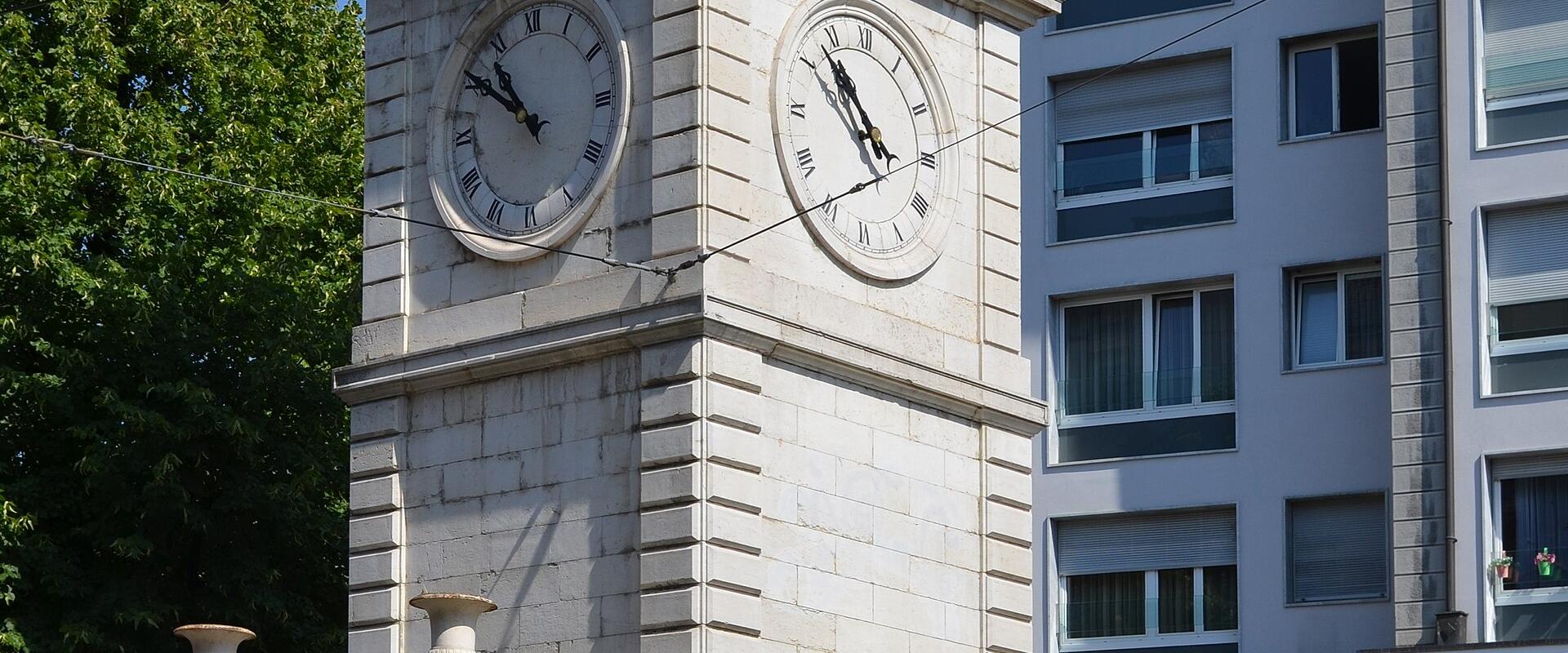 A stone clock tower with a square base, two visible clock faces with Roman numerals, and a louvered belfry topped by a pediment, situated next to a modern apartment building.