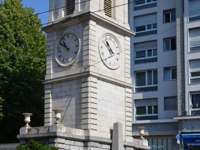 A stone clock tower with a square base, two visible clock faces with Roman numerals, and a louvered belfry topped by a pediment, situated next to a modern apartment building.