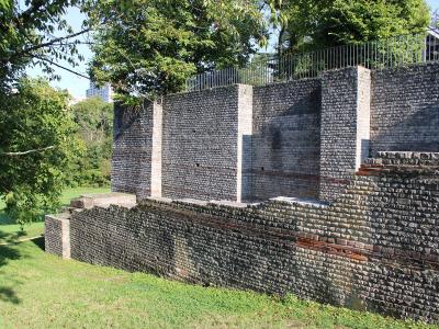 A section of ancient Roman brick walls and foundations set within a grassy archaeological park, with modern trees and a building visible in the background.