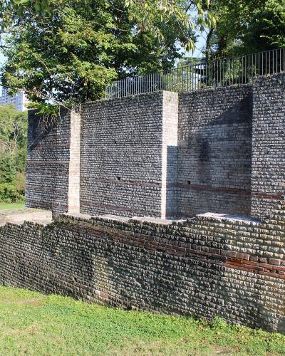 A section of ancient Roman brick walls and foundations set within a grassy archaeological park, with modern trees and a building visible in the background.