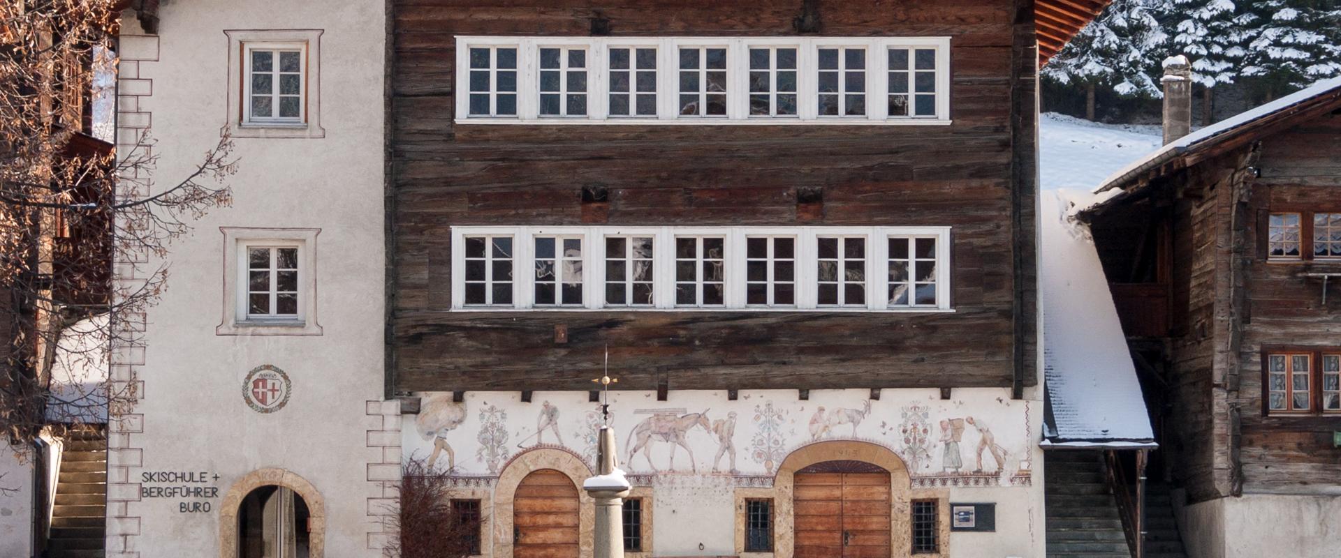 A large, traditional Swiss building with a white stucco left section and a dark wooden upper section, featuring murals on the ground floor and text reading "SKISCHULE + BERGFÜHRER BÜRO" on the wall, situated in a snowy village square with a fountain 