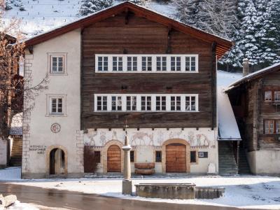 A large, traditional Swiss building with a white stucco left section and a dark wooden upper section, featuring murals on the ground floor and text reading "SKISCHULE + BERGFÜHRER BÜRO" on the wall, situated in a snowy village square with a fountain 
