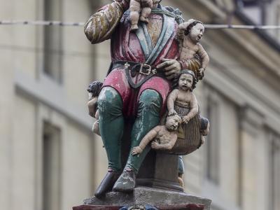 A close-up photograph of a painted stone statue depicting a grotesque figure with a pointed hat eating a small child, while holding other children, mounted on an ornate fountain column with a classical building facade in the background.