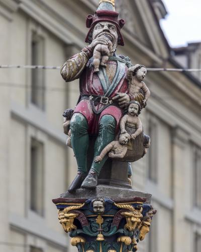 A close-up photograph of a painted stone statue depicting a grotesque figure with a pointed hat eating a small child, while holding other children, mounted on an ornate fountain column with a classical building facade in the background.