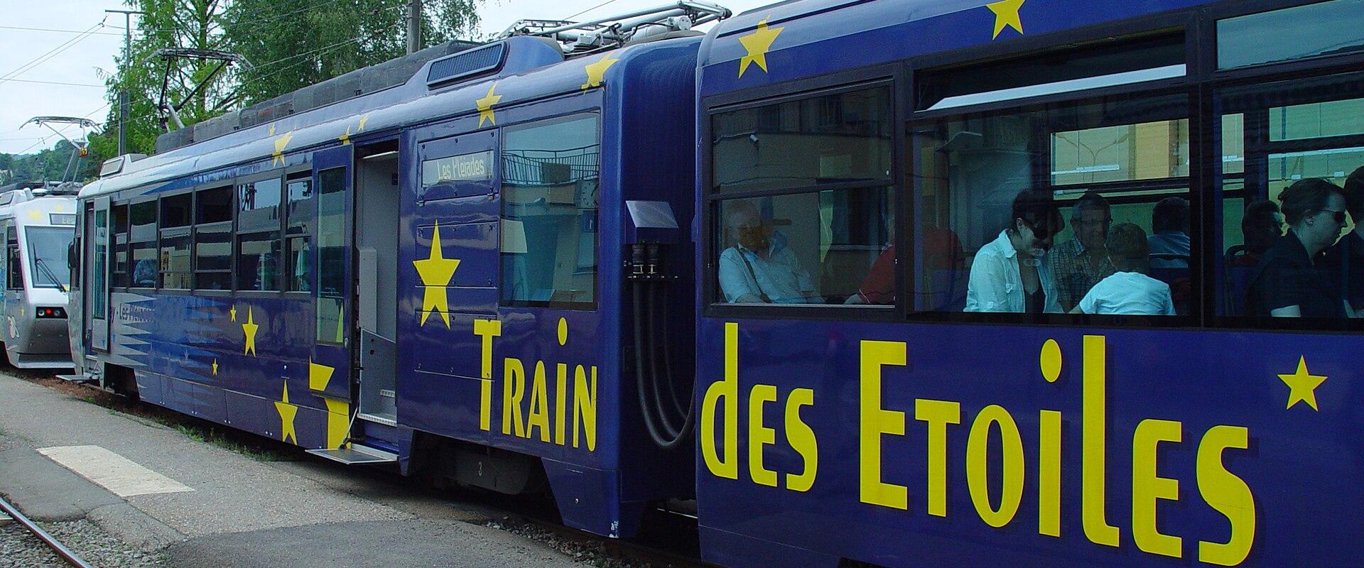 A blue cogwheel train carriage decorated with yellow stars and the text "Train des Etoiles" is stopped at a station platform with passengers visible inside.