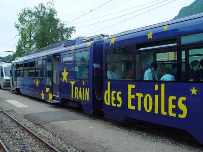A blue cogwheel train carriage decorated with yellow stars and the text "Train des Etoiles" is stopped at a station platform with passengers visible inside.