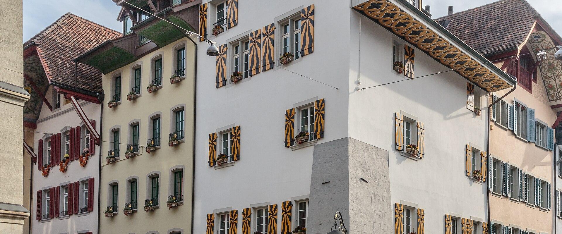 A street-level view of a white corner building with distinctive yellow and black striped shutters and the word "LATERNE" on the facade, located on a cobblestone street with other historic buildings.